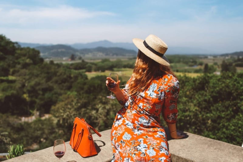 Woman in red floral dress and straw hat drinking wine on a ledge at a Napa Valley Winery