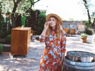 Woman in red floral dress and straw hat drinking wine next to a barrel at Grgich Hills Estate