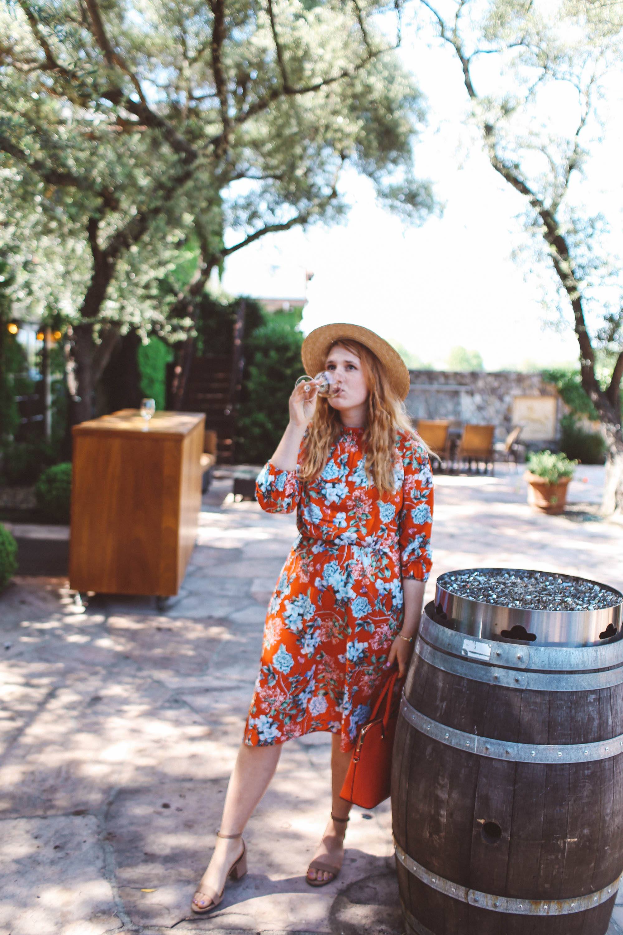 Woman in red floral dress and straw hat drinking wine next to a barrel at Grgich Hills Estate