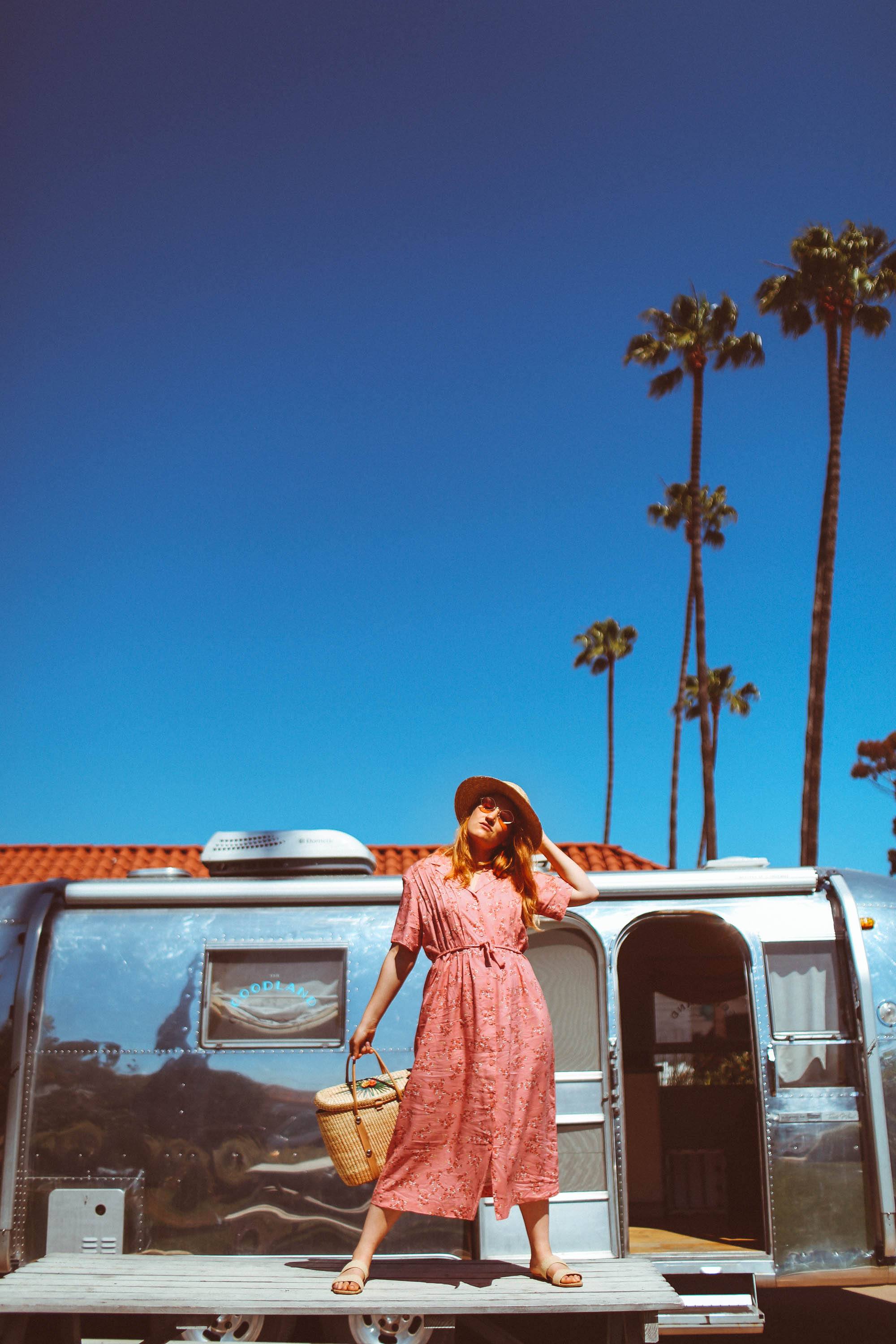Woman in hat and vintage pink dress and hat standing on a table in front of an airstream with palm trees in the background