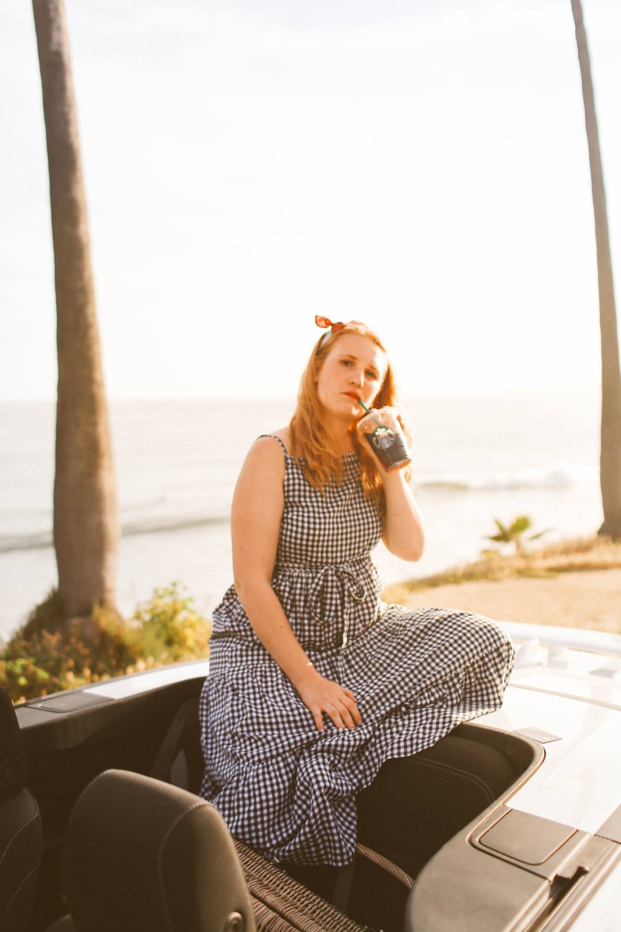 Woman in black and white gingham dress sitting on convertible drinking starbucks