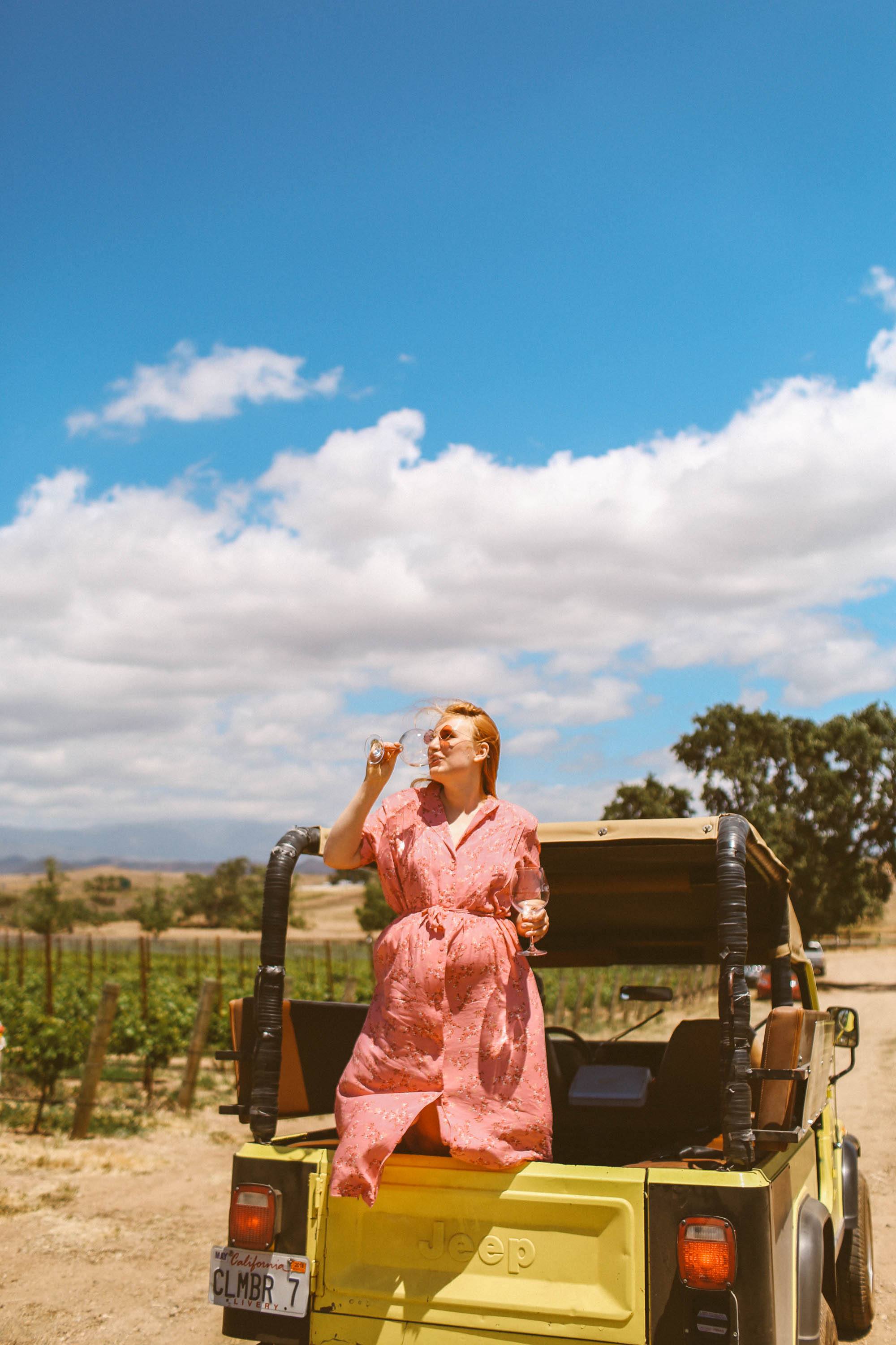 Woman in vintage pink dress drinking wine on the back of a jeep at Gainey vineyards