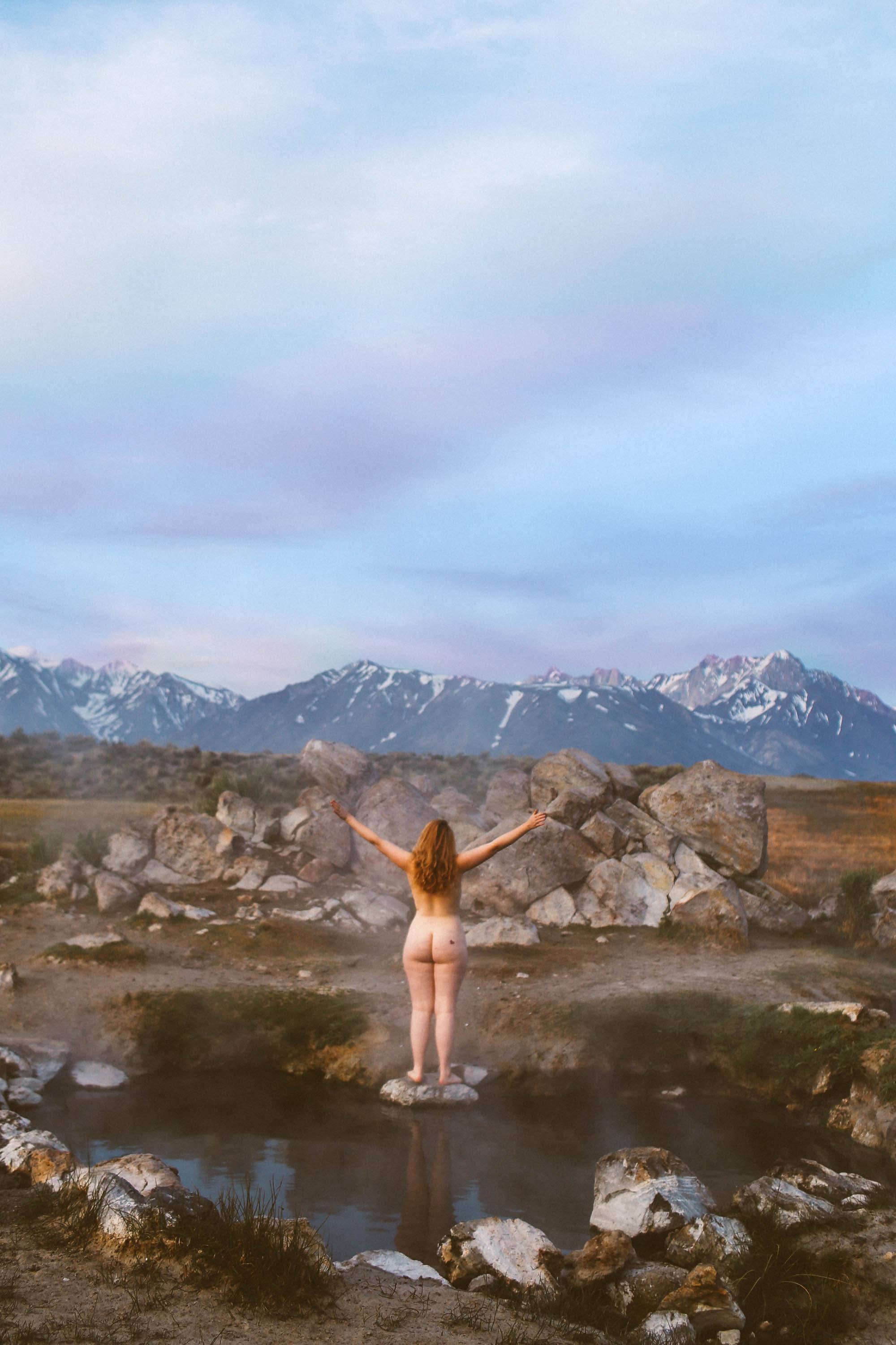 Woman standing naked on rock in a hot spring with snow capped mountains in the background